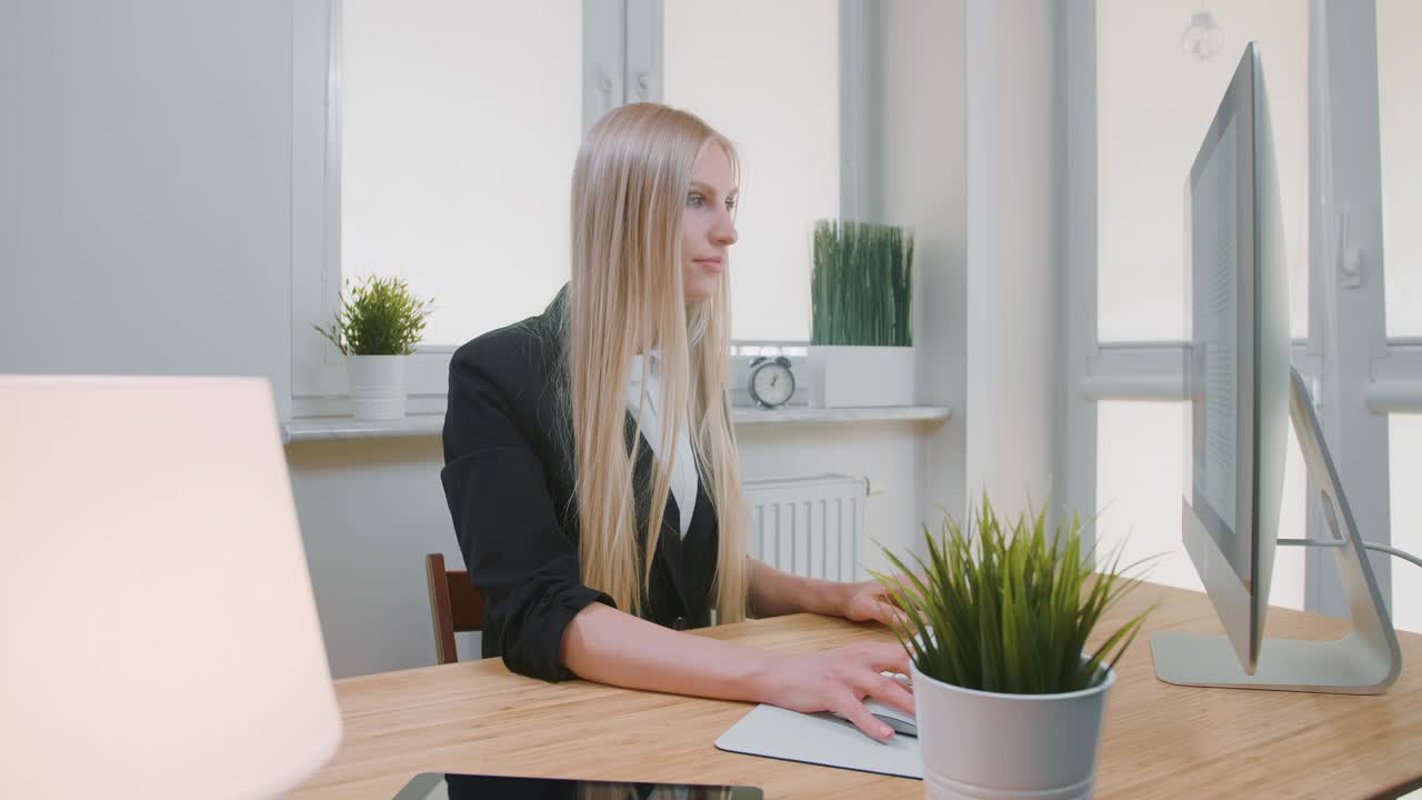 Donna sorridente che lavora al computer in ufficio. Bella giovane donna bionda seduta alla postazione di lavoro con il computer e con la mano sul mouse, che guarda la telecamera e sorride