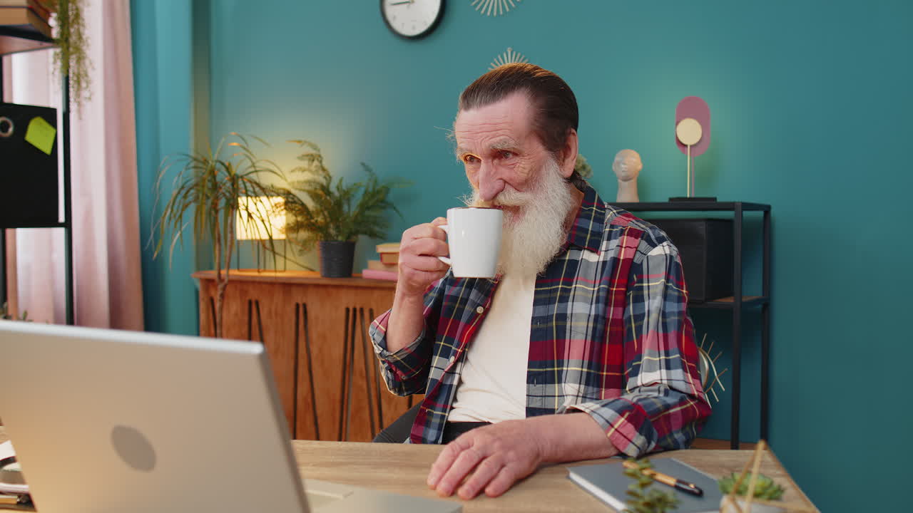 Elderly businessman working on laptop computer drinking cup of warm hot coffee sits at home table
