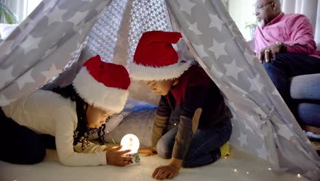 african american brother and sister in christmas hats using snow globe in blanket tent, slow motion