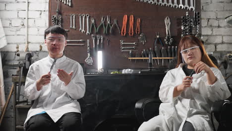 chinese lab technicians seated in automotive workshop, boy tapping vernier caliper on hand while wearing safety goggles, lady holding screw, industrial workspace with tools equipment on wall