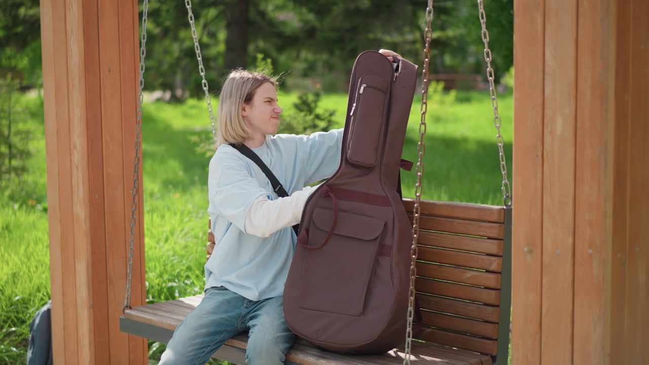 chitarrista caucasico che accorda la chitarra su una panchina di legno oscillante sotto un gazebo, musicista biondo che rimuove la chitarra dalla custodia imbottita, regola la tracolla e le corde, espressione concentrata, abbigliamento casual con felpa di jeans, estate