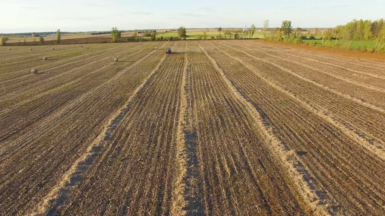 Weitwinkelansicht eines staubigen braunen Ackers, während ein Traktor eine Ballenpresse zieht, um große Rundballen für den Winter zu pressen und einzulagern.