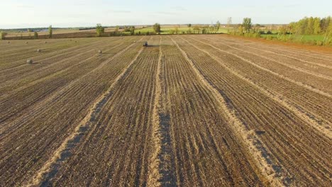 vista panorámica de un campo agrícola marrón polvoriento mientras un tractor remolca una máquina empacadora para hacer grandes fardos redondos que se almacenarán para su uso en invierno