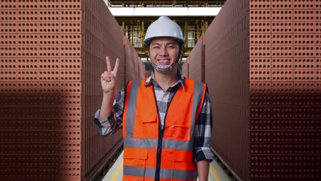 asian male engineer with safety helmet smiling and showing peace gesture while standing with red brick packed in stacks are stored