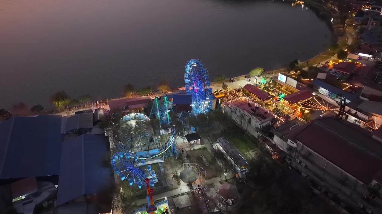 Toma aérea con dron de una colorida noria iluminada y atracciones de parque de atracciones junto a la orilla del lago por la noche