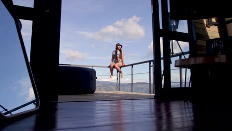 Wide-shot-of-a-woman-in-bathing-suit-seated-on-a-fence-with-the-horizon,-the-say-and-clouds-behind-her