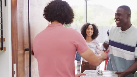 Happy-biracial-man-greeting-diverse-friends-with-flowers-and-christmas-present-at-his-front-door