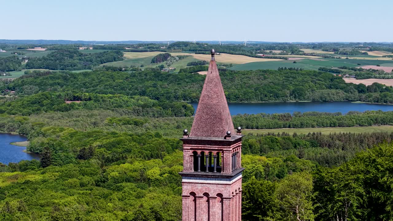 Drone footage featuring a red-brick tower with a pointed spire rising above lush green forest, with farmland fields and a lake in the background, offering panoramic countryside views in Denmark