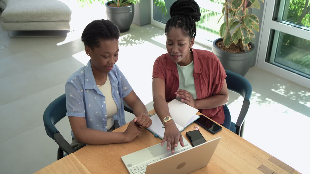 African American female friends leaning in right woman pointing at laptop guiding left woman typing