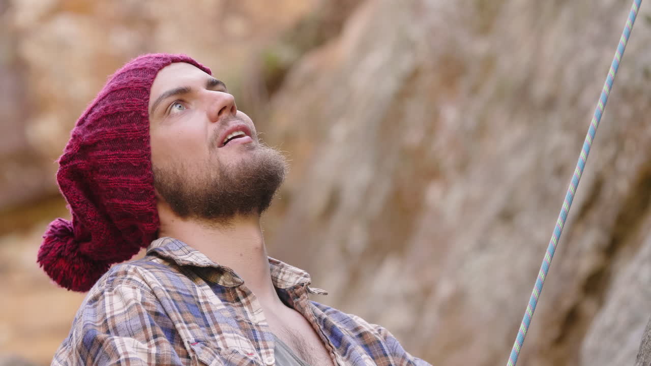 Medium close up of bearded male mountaineer wearing flannel shirt and red beanie belaying a rope while looking up and talking with his rock climbing partner
