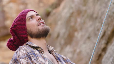 medium close up of bearded male mountaineer wearing flannel shirt and red beanie belaying a rope while looking up and talking with his rock climbing partner