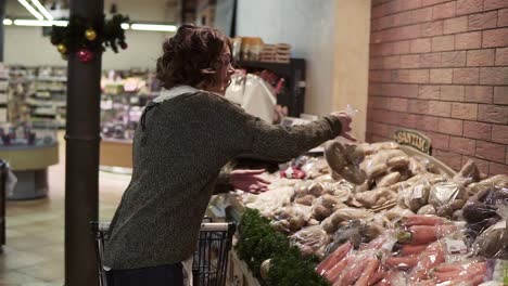 una mujer caucásica está conduciendo un carrito de compras a través del departamento de alimentos en un supermercado y recogiendo dos bolsas con patatas.