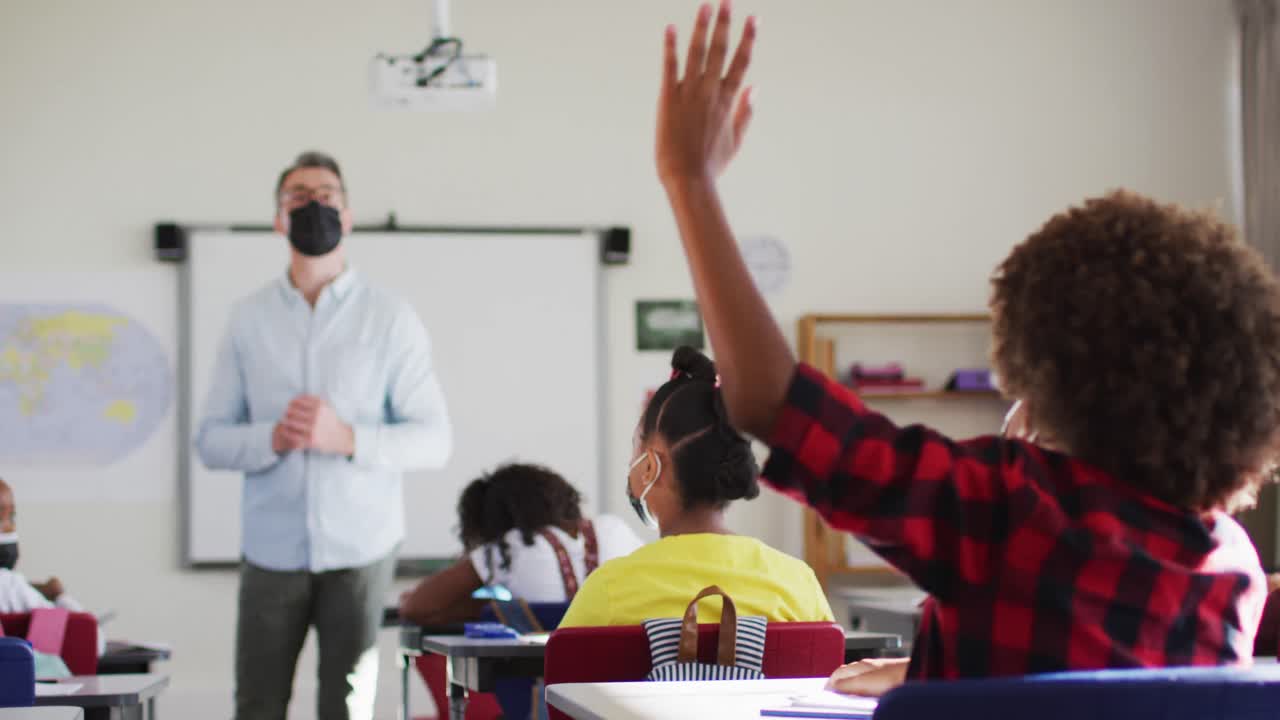 maestro masculino diverso en el aula con niños levantando las manos durante la lección, todos con máscaras faciales