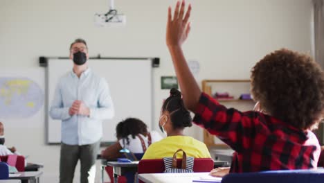 diverse male teacher in classroom with children raising hands during lesson, all wearing face masks