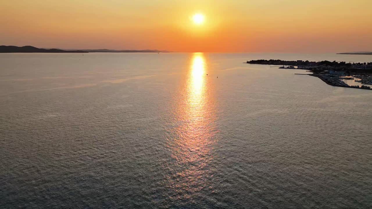 Expansive drone view over a coastal cityscape at sunset. The orange sun dips low over the Adriatic, casting glowing reflections across calm, peaceful sea waters. A serene aerial moment, Zadar, Croatia