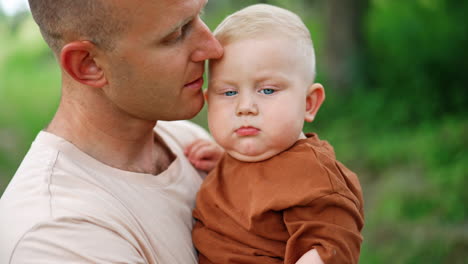 man holds a baby boy and kisses him on the head. father and son portrait close up in the nature.