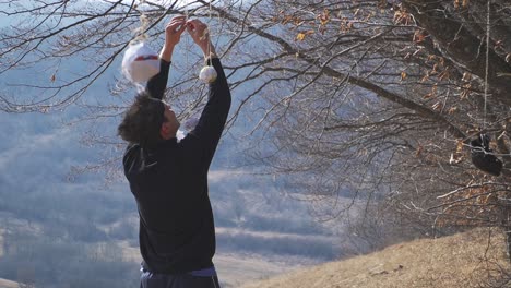 a ninja in black attire carefully sets up hanging targets on a tree, preparing for precision training. the scenic countryside setting enhances the atmosphere of discipline and martial arts mastery