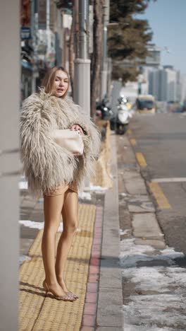 a stylish woman stands on a snowy sidewalk looking out for a taxi wearing a shaggy fur coat, beige tights, and heels, holding a white bag, with an urban background of buildings and vehicles in seoul