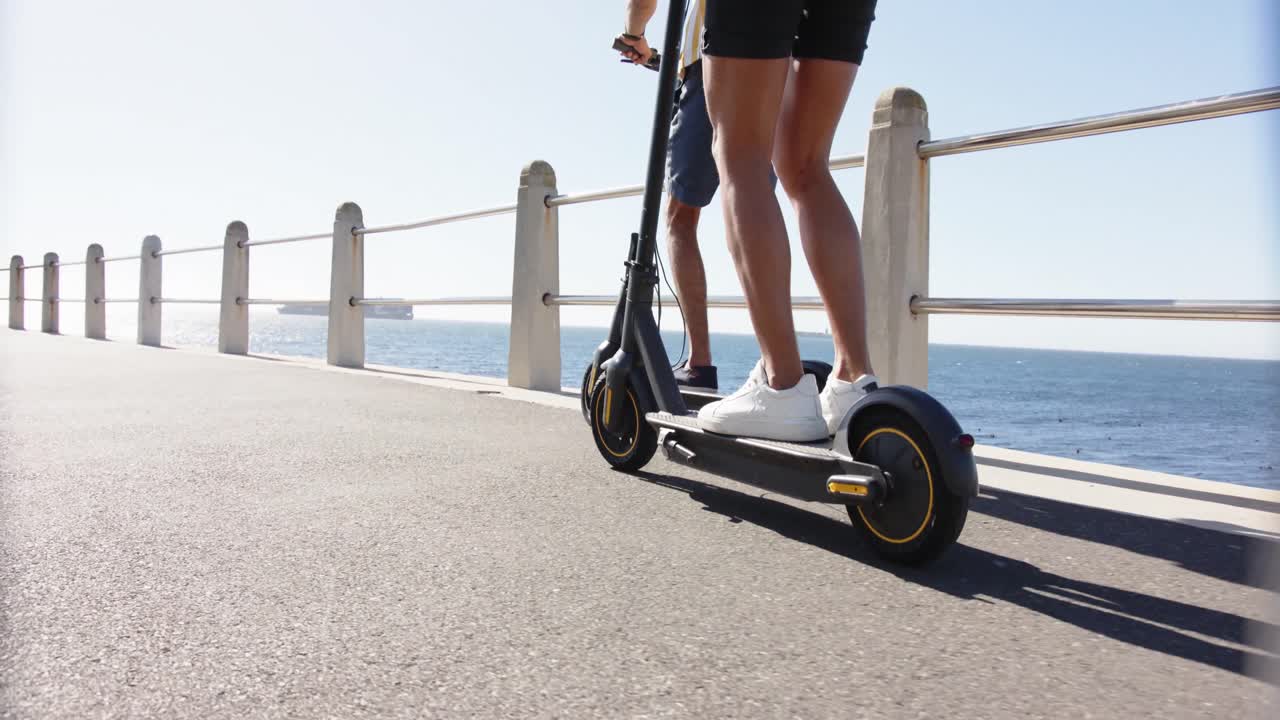 Midsection of diverse gay male couple using scooters at promenade by the sea, slow motion