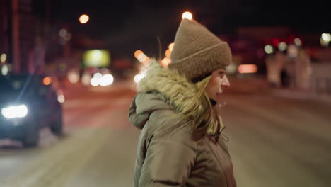 first-person view of a woman in a winter coat with a fur hood, crossing the road at night with blurry people and city lights in the background