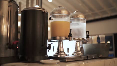 detailed shot of orange juice and water coolers at breakfast drinks table in hotel dining room, with white granite table underneath and other large coolers