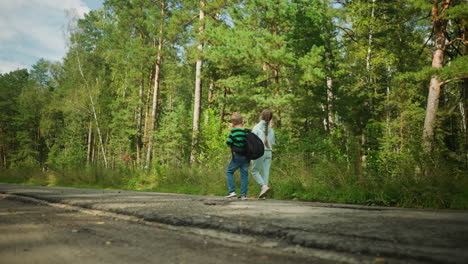 close up of cracked tar road foreground with siblings walking along roadside in lush green forest, boy carrying backpack and girl strolling beside uneven pavement under sunlit woodland canopy