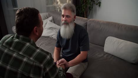 Over-the-shoulder-A-man-with-gray-hair-and-a-lush-gray-beard-in-a-gray-T-shirt-communicates-and-holds-the-hand-of-his-boyfriend-a-brunette-with-stubble-in-a-checkered-shirt-sitting-on-a-gray-sofa-in-the-evening-in-a-modern-apartment