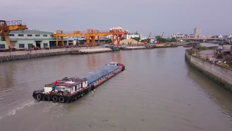 Boat-Transporting-Goods-making-its-way-along-Bang-River-between-Industrial-Sites-and-Loading-Cranes,-Shanghai,-China