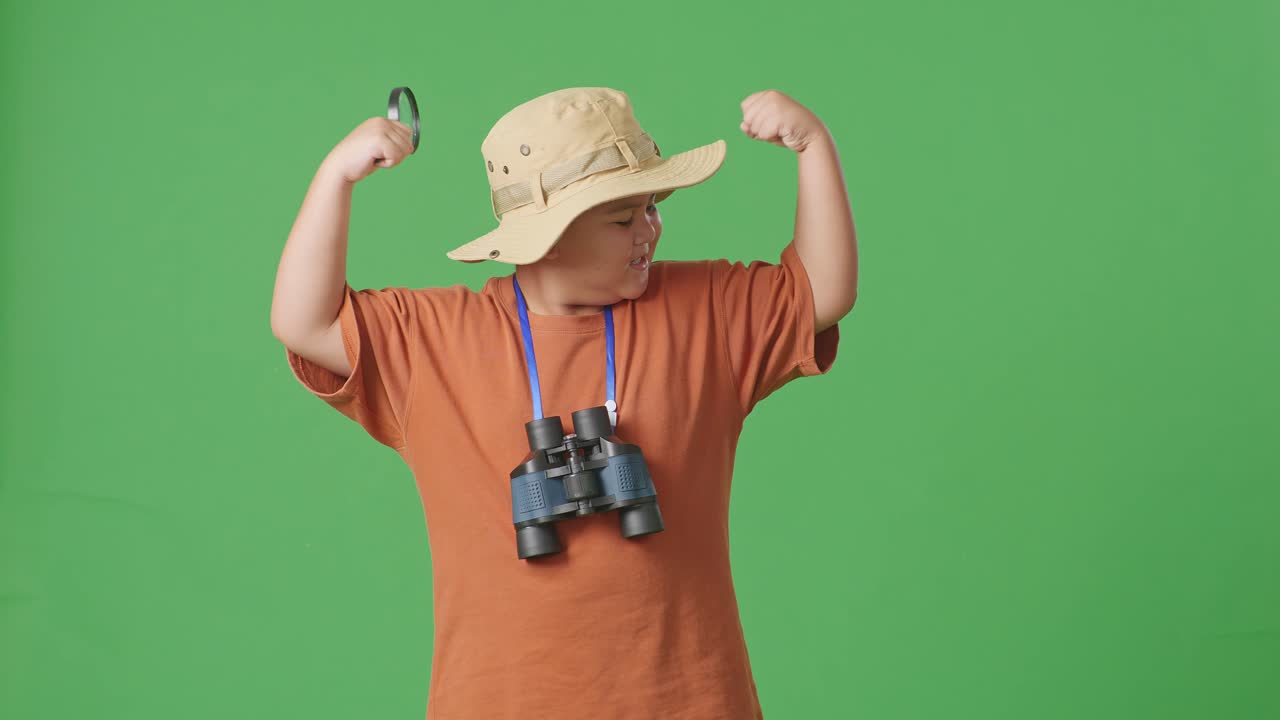 모자와 안경을 쓴 아시아 관광 소년은 보기를 사용하고 녹색 스크린 배경에 이를 구부린다. 소년 연구자는 여행 관광 모험 개념의 무언가를 조사합니다.