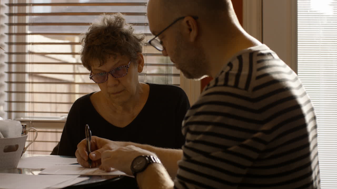 An elderly woman sits at a kitchen table with a younger man. He is her adult son, who's also her caregiver post stroke. They are reviewing, and signing documents together. Both wear reading glasses.