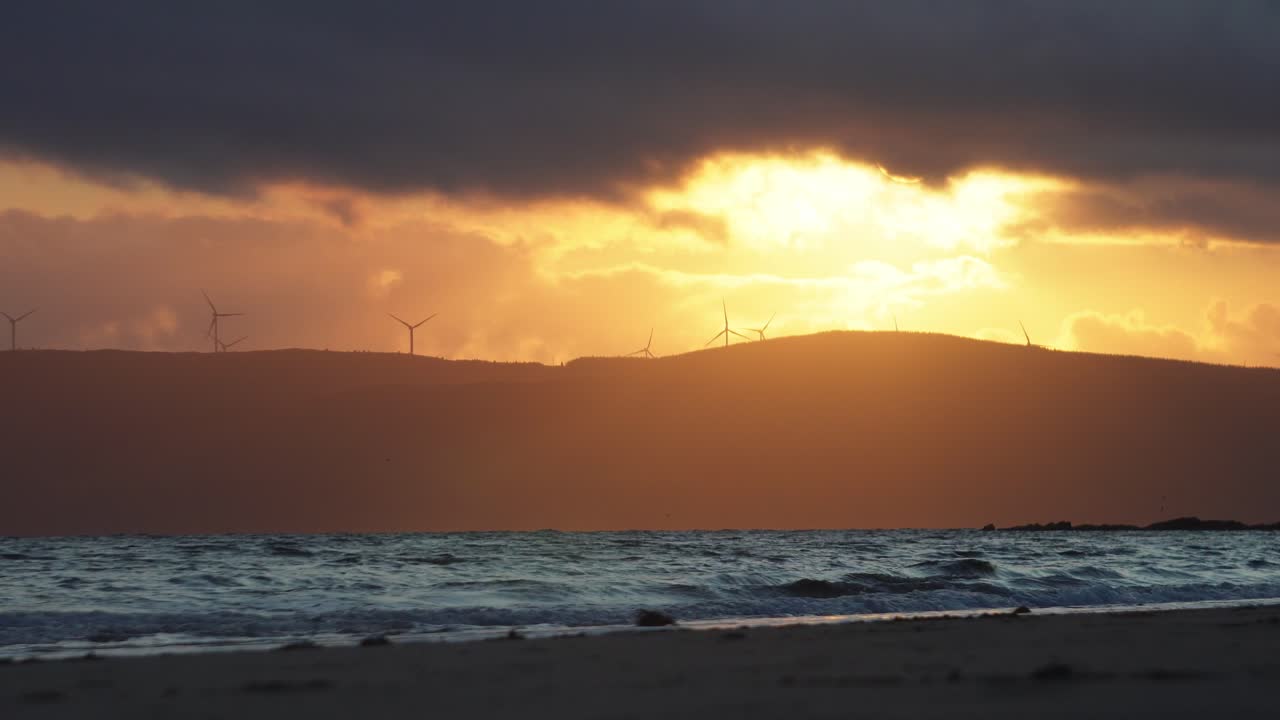 A scenic sunset over a Scottish bay with waves crashing onto shore, silhouetted mountains in the distance, and wind turbines turning on the ridgeline. A calm, atmospheric coastal scene.