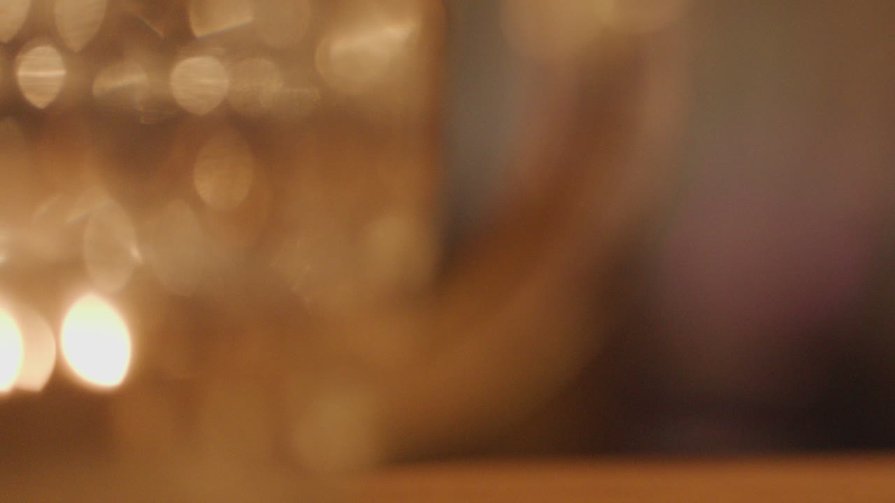 A hand reaches for a whisky glass on a wooden bar, surrounded by blurred glasses and warm candlelight, with shallow focus and soft bokeh