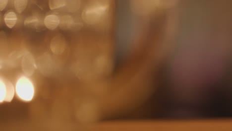 a hand reaches for a whisky glass on a wooden bar, surrounded by blurred glasses and warm candlelight, with shallow focus and soft bokeh