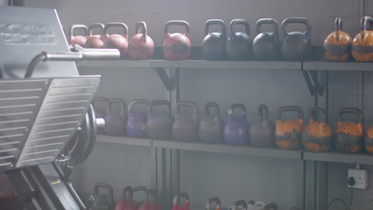 La telecamera sta panoramando a destra, rivelando kettlebell arancioni su un ripiano e una presa, mentre la macchina a sinistra rimane ferma