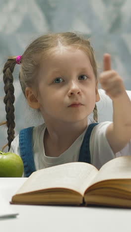 slow motion portrait of little child student during home schooling and self-isolation, which pauses, eats green mouthwatering apple, shows thumbs up means class, with tutorial and notebooks on table