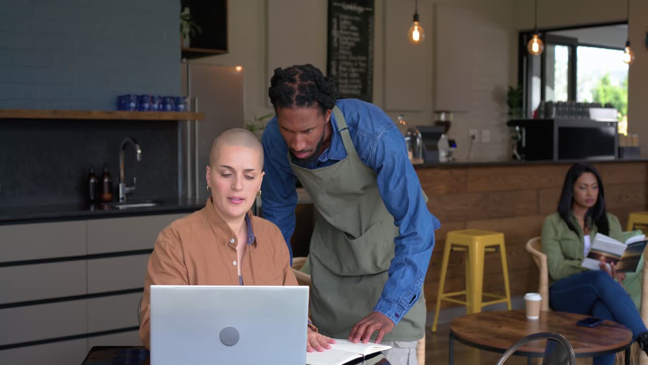 Zwaaiend voor de aandacht van de barista typt een vrouw notities op haar laptop aan een cafétafel, terwijl de barista eraan komt.