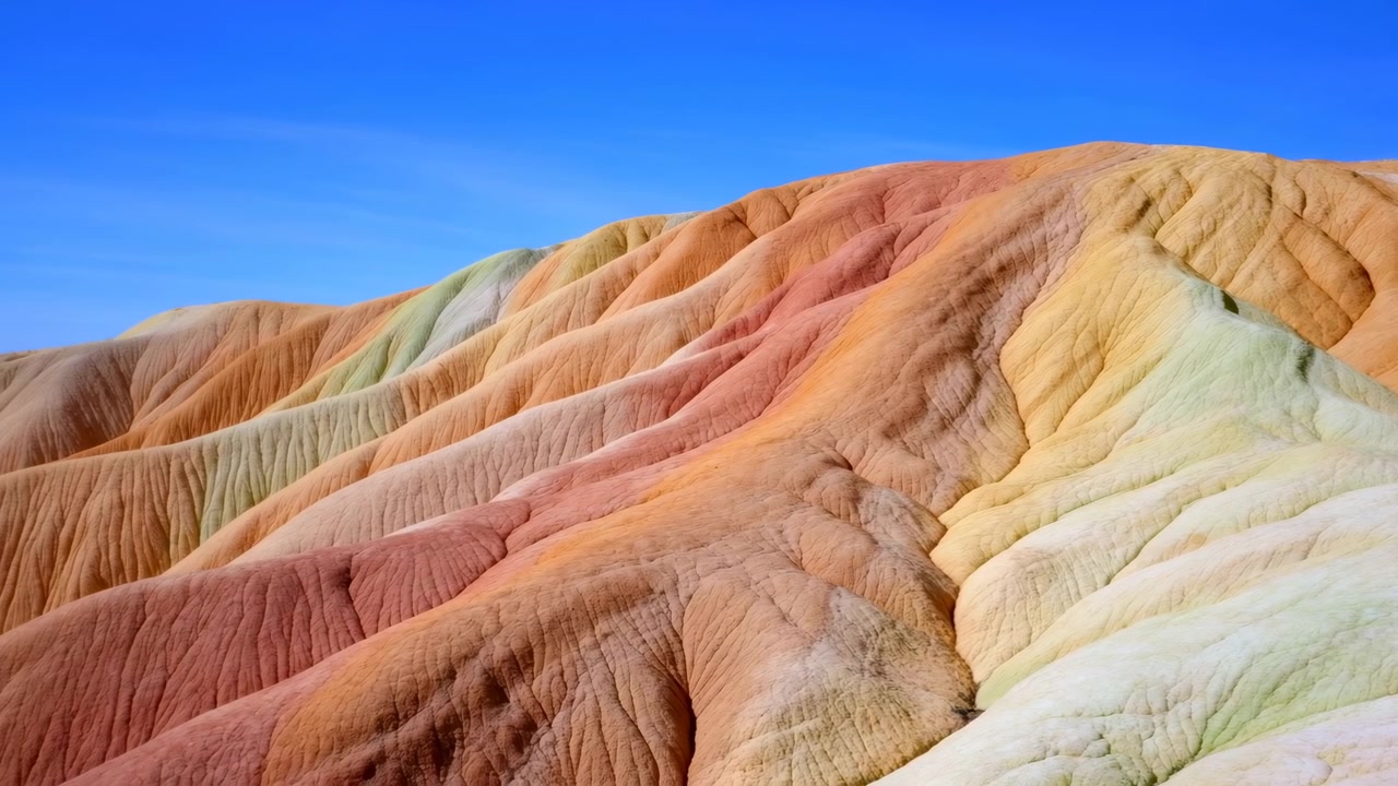 Cume colorido e estratificado de badlands, com colinas de argila lisas e erodidas sob um céu azul límpido. A vista, de um ângulo baixo e amplo, revela tons minerais pastel, textura desgastada, formas arredondadas e uma vasta imensidão.