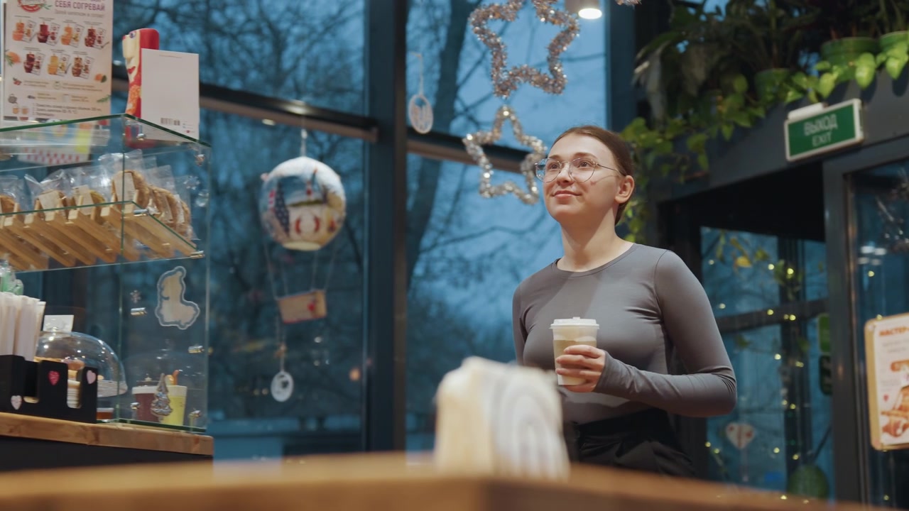 Donna asiatica che ammira le decorazioni e le luci natalizie del caffè mentre tiene in mano un caffè, guardando in alto gli ornamenti appesi e i riflessi scintillanti, top grigio casual e grembiule visibili, interni serali accoglienti