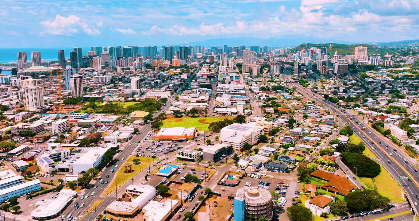 Quang cảnh từ trên không mạng lưới đô thị Honolulu và đường chân trời trung tâm thành phố, Hawaii. Mạng lưới đường phố, khu dân cư và thương mại hỗn hợp, đại dương và những ngọn đồi xanh ở phía xa dưới bầu trời nhiều mây một phần