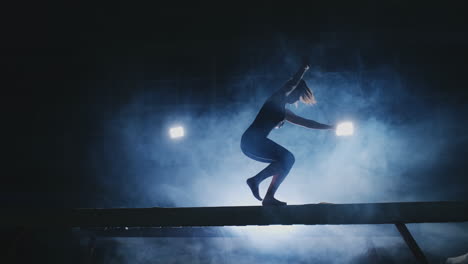 la niña realiza un truco en un tronco con luz de fondo y cámara lenta en ropa de gimnasia deportiva. humo y azul. saltar y girar en la viga de equilibrio