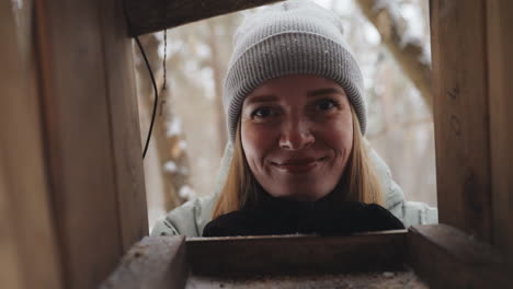 woman in gray beanie and winter jacket smiling warmly while peering into wooden bird feeder in snowy forest, hands together, peaceful expression, enjoying quiet moment in nature during cold season