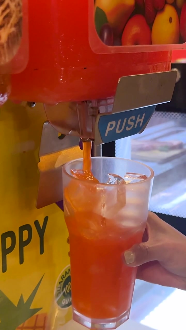 En hånd holder en kop is under en dispenser, mens appelsinjuice hældes ud