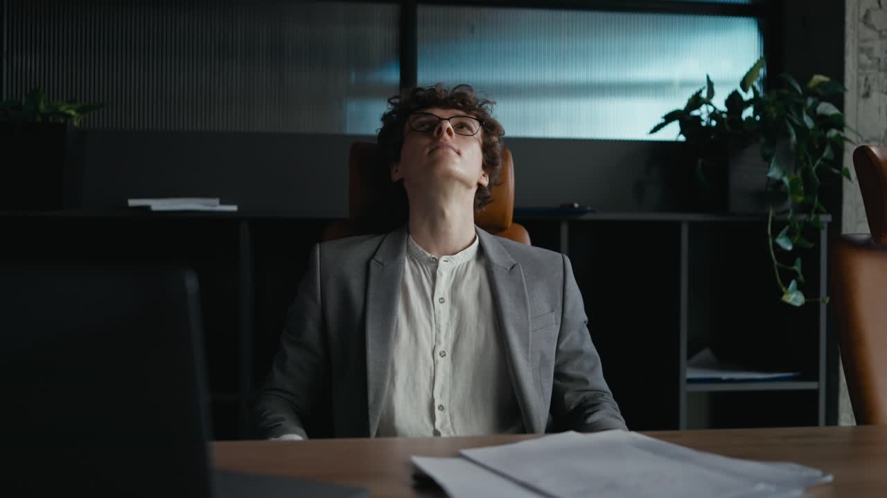 Confident young guy with curly hair in a gray jacket examines papers and leans back in his chair to relax while working in the office