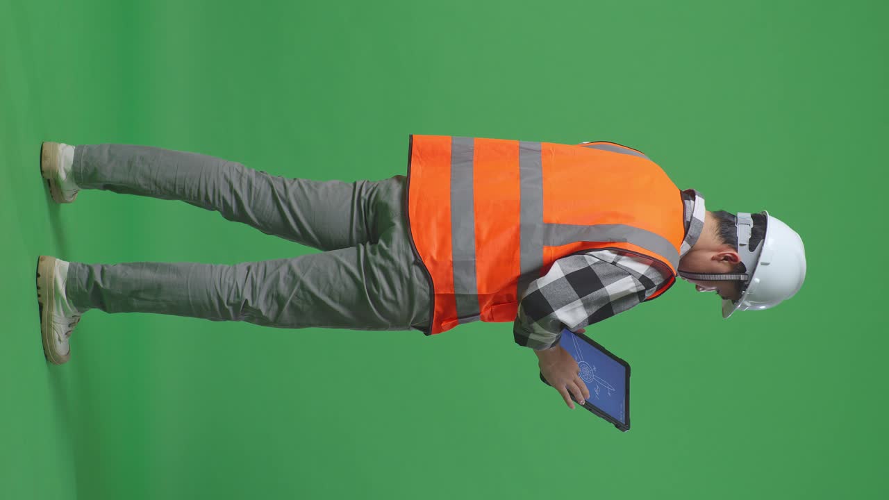 Full Body Back View Of Asian Male Engineer With Safety Helmet Looking At The Wind Turbine Blueprint On A Tablet While Standing In The Green Screen Background Studio