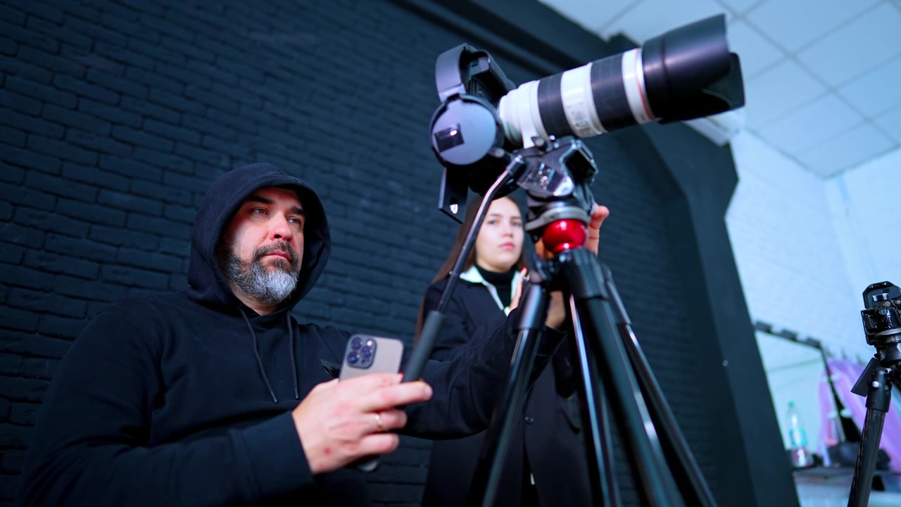 Videografo che Regola la Telecamera su Treppiede con Assistente di Studio. Uomo con Felpa Nera Regola la Telecamera su Treppiede con Obiettivo Teleobiettivo mentre l'Assistente è in Piedi Vicino a un Muro di Mattoni Neri.