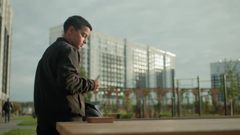 student retrieves study material from backpack and places it on wooden table in outdoor setting with blurred background of tall buildings and natural light creating calm academic atmosphere