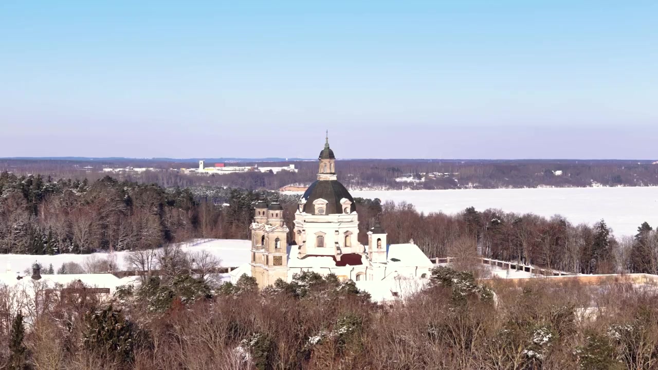 Paesaggio invernale con bosco e una torre di monastero in lontananza sotto un cielo azzurro e limpido.