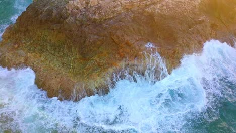 tilting up aerial shot of foamy waves hitting a cliff at sunset