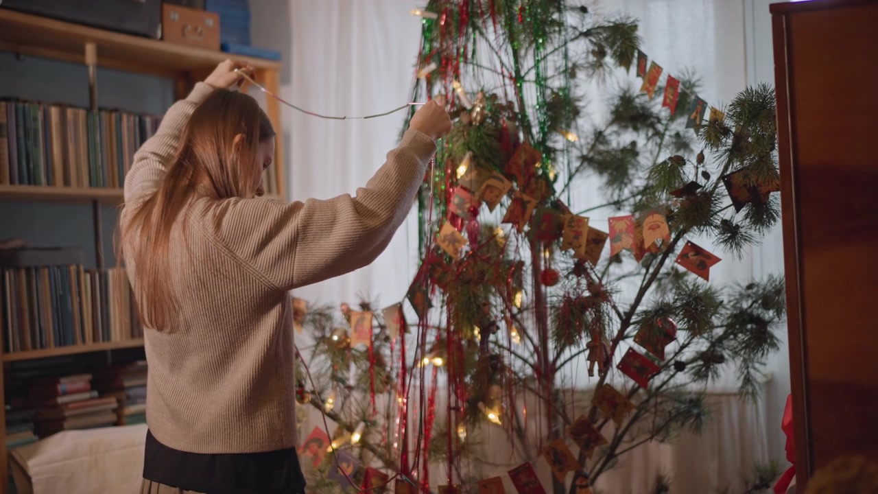White woman strings lights on studio tree by bookshelf and records, arranging handmade ribbons and paper cards indie musician ritual in warm evening light preparing seasonal display with creative
