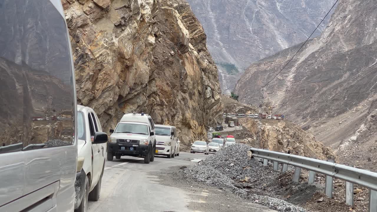 Line of cars cautiously driving along Gilgit Skardu Road, navigating past roadside rubble from a recent landslide. A challenging journey through a rugged mountainous terrain.